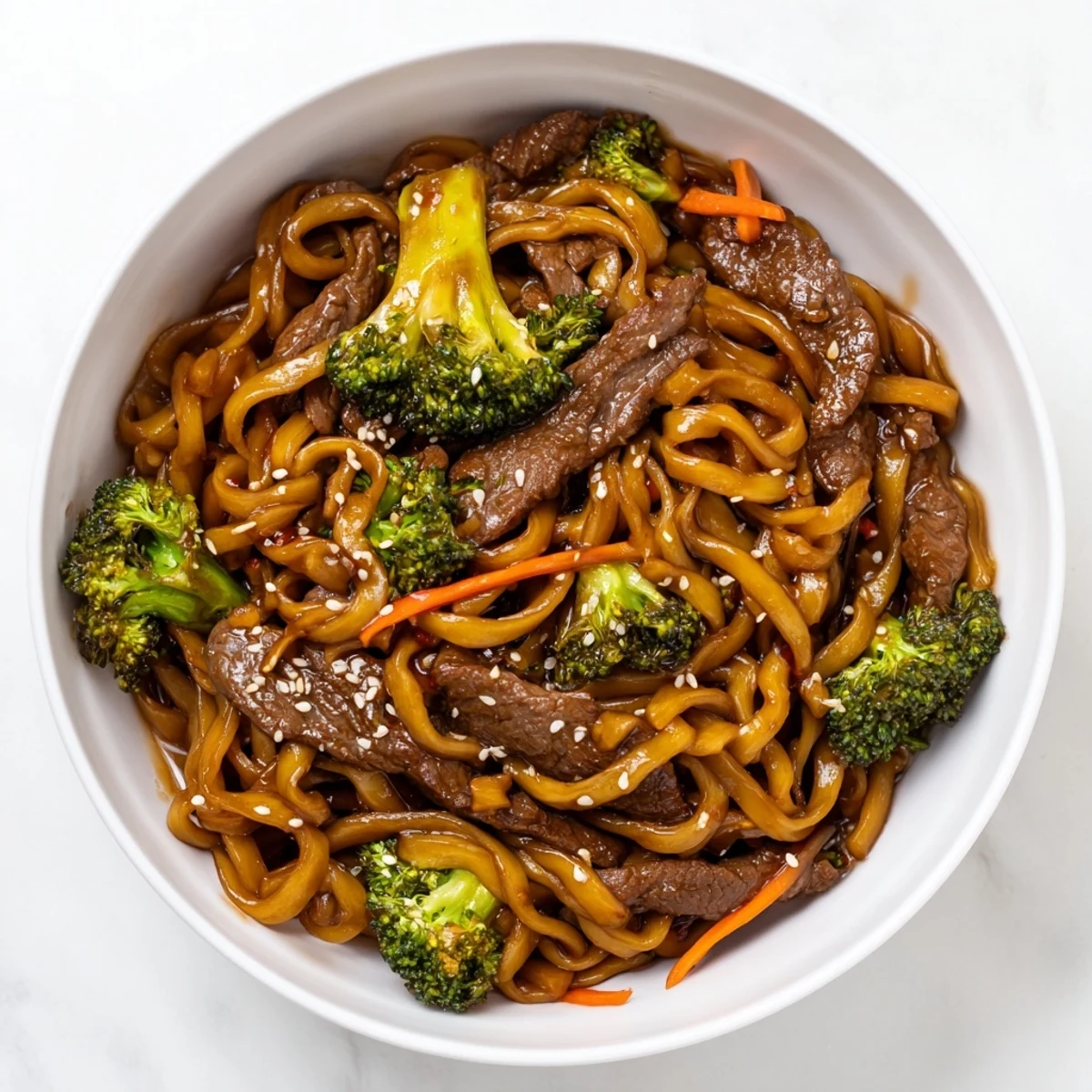 Tender beef and silky noodles are tossed with bright green broccoli in a rich soy glaze, served hot and garnished with sesame seeds on a ceramic plate.