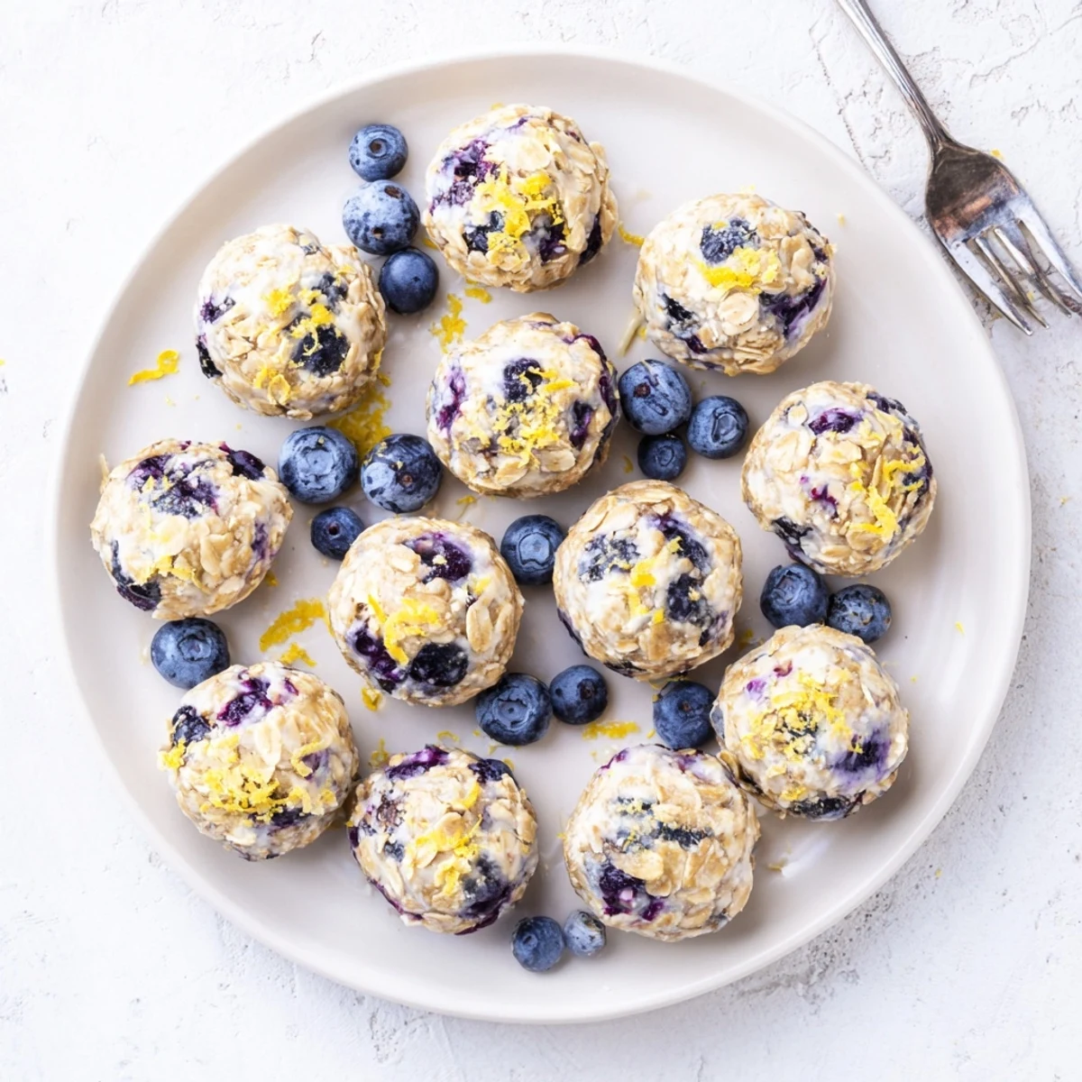 Five blueberry cheesecake protein bites arranged on a white marble surface beside a glass of milk and fresh blueberries for a wholesome snack serving idea.