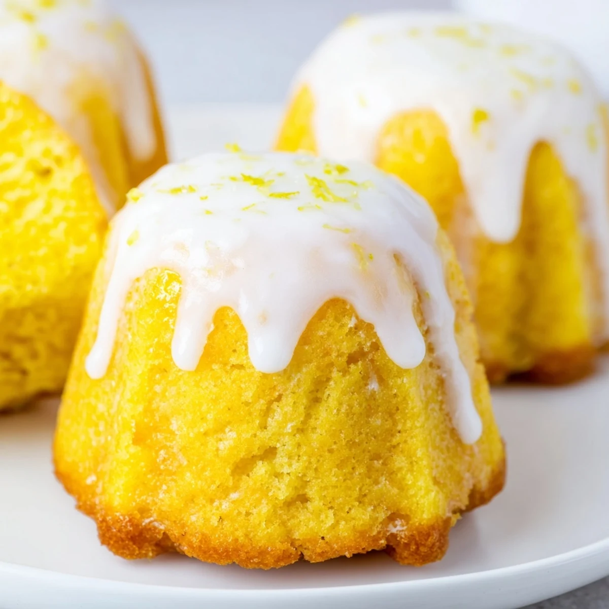 Golden mini lemon cakes with drizzled glaze sit on a white serving plate